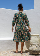 Model stands backwards in cotton tiered over the knee floral print dress, holding the skirt out, paired with tan sandals