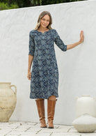Woman wearing a cotton navy blue based floral dress with sleeves standing against a white wall with decorative pots.