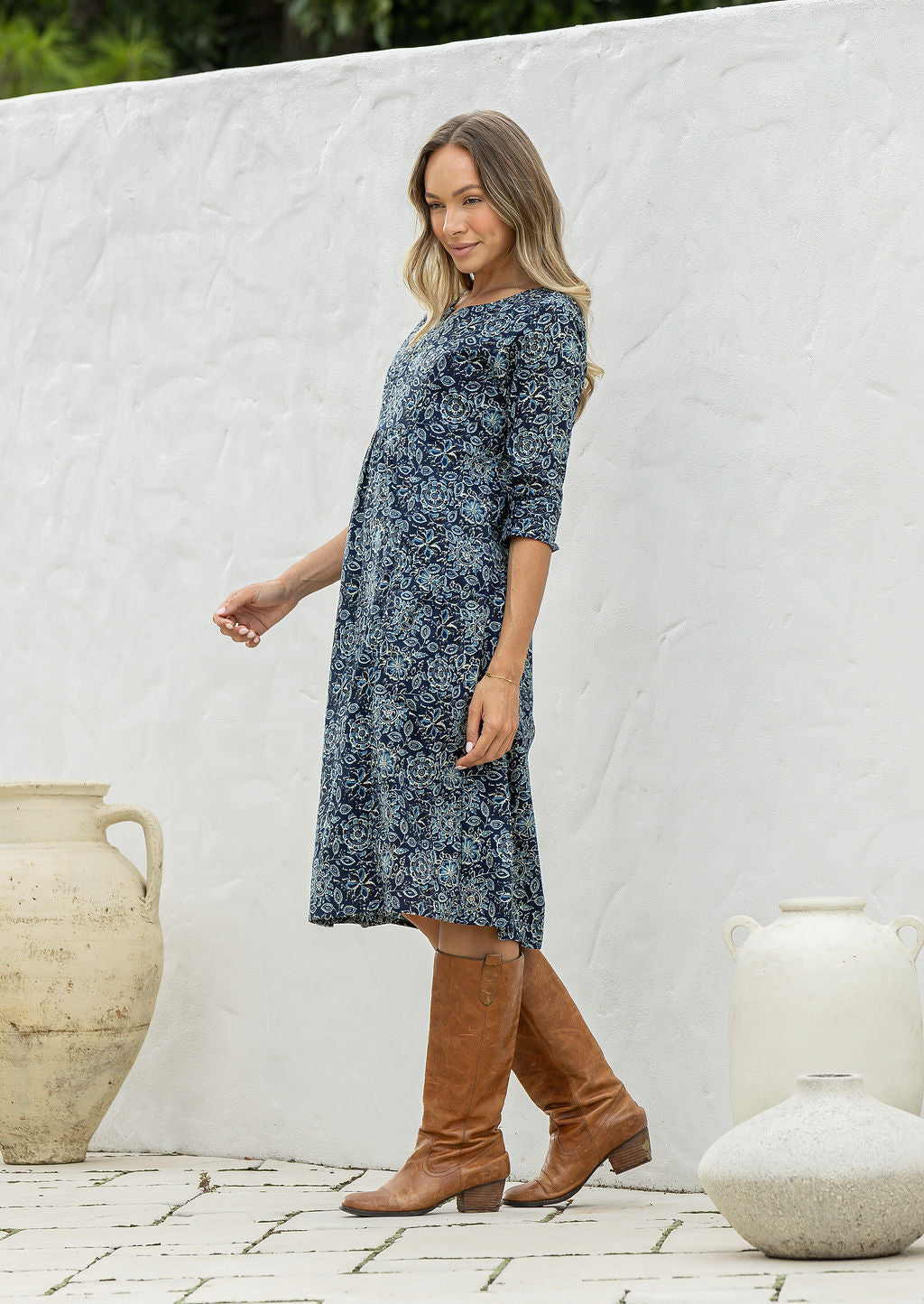 Woman stands sideways wearing a cotton navy blue based floral dress with sleeves standing against a white wall