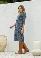 Woman stands sideways wearing a cotton navy blue based floral dress with sleeves standing against a white wall