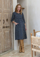 Woman poses in a 100% cotton navy blue based floral dress with sleeves and one hand in pocket