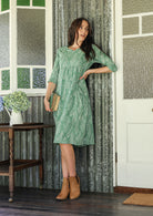 Woman wearing a cotton sage green based floral dress with sleeves standing in a rustic room