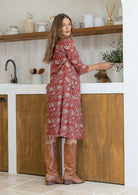 Woman stands sideways wearing a cotton red based floral dress with sleeves and knee-high boots standing in a kitchen.