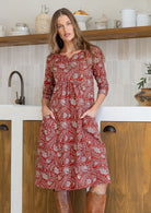 Woman wearing a 100% cotton red based floral dress with sleeves and hands in pocket.