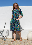 A woman modelling a knee-length cotton dress with a vibrant multicolour floral and peacock print on a dark navy background, standing with hands in pockets