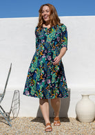 A woman wearing a knee-length floral peacock print cotton dress with three-quarter sleeves stands outdoors against a white wall under a blue sky