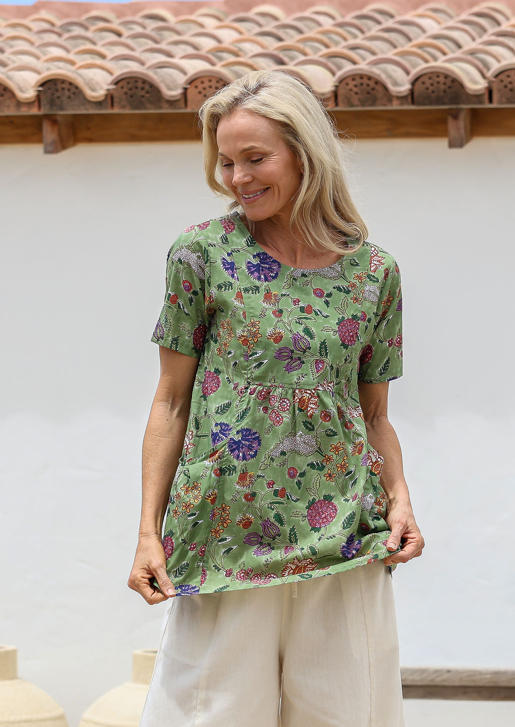 Woman wearing a moss green floral top with round neckline, short sleeves and pockets with pin tucks, standing against a white wall with a terracotta roof.