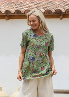 Woman wearing a moss green floral top with round neckline, short sleeves and pockets with pin tucks, standing against a white wall with a terracotta roof.