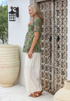 Woman in a green floral top with pockets and white pants standing next to a rustic wooden door and a large beige pot.