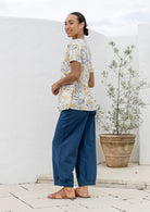 Woman stands sideways in a cotton neutral-based top featuring an empire waist, short sleeves and pockets.