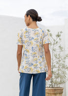 Woman standing backwards wearing a  short sleeved neutral based floral cotton top with empire waistline and deep pockets, paired with plain blue cotton pants