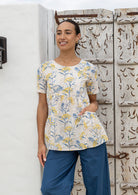 Woman wearing a neutral based floral print top with short sleeves, an empire waistline and deep pockets, standing against a wooden door