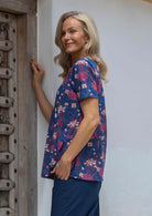 Woman stands sideways in a cotton blue-based top with pink, plum, and burgundy floral print, featuring an empire waist, short sleeves and pockets.