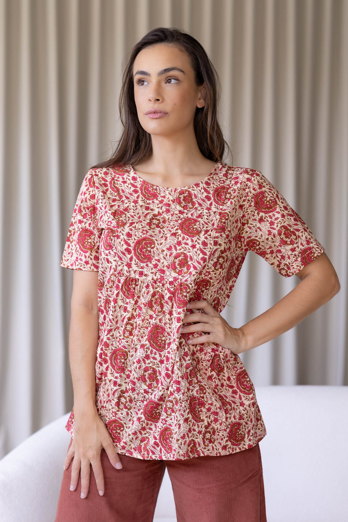 Woman standing wearing a 100% cotton short sleeved gentle cream based floral cotton top with empire waistline and deep pockets, paired with plain pants