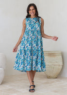 Woman wears cotton sleeveless three tiered midi length dress in aqua and royal blue daisy print