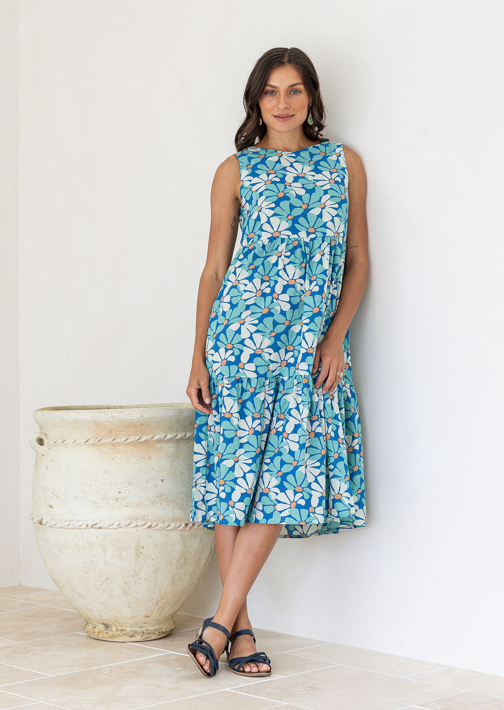 Woman leans on wall wearing a cotton sleeveless three tiered midi length dress in aqua and royal blue daisy print