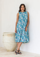 Woman leans on wall wearing a cotton sleeveless three tiered midi length dress in aqua and royal blue daisy print