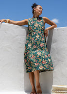 Woman in a sleeveless, floral green based 100% cotton dress standing in front of white wall. 