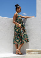 Woman stands in side pose wearing a tiered sleeveless sundress with pockets, high round neck and empire waist.