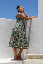 Woman standing against a white wall wears relaxed floral cotton dress with green based floral print.