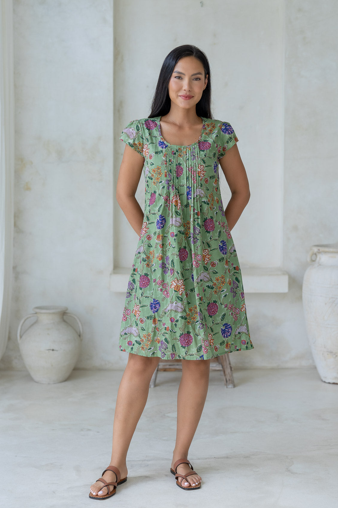 Woman wears cotton cap sleeved above knee dress with round neckline and small pleats across the bodice, in a moss green based folk art style floral print.