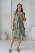 Woman wearing a moss green floral cotton dress with cap sleeves, round neckline and small pleated design across the bodice.