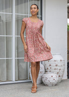 Woman wearing a 100% cotton rose pink based floral dress with round neckline and cap sleeves standing in front of decorative vases.