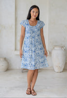 Woman wearing a cap sleeved, above knee length, white based cotton dress with blue floral print and lemon yellow highlights.