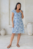 Woman wearing a white floral cotton dress sitting above knee length with pleats across the bodice, standing in a room with white walls and large vases.