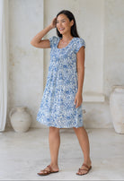 Woman wearing lightweight cotton dress with round neckline, small pleats across the bodice and cap sleeves in white based blue floral print.