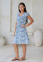 Woman posing in 100% cotton above knee length dress with round neckline, cap sleeves and small pleats across the bodice in white based floral print.