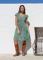 Woman wearing a 100% cotton sage green based floral dress standing in front of a white wall. 