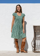 Woman wearing a green floral dress and brown boots standing against a white wall.