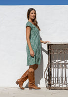 Woman stands sideways wearing a 100% cotton sage green based floral dress with round neckline.