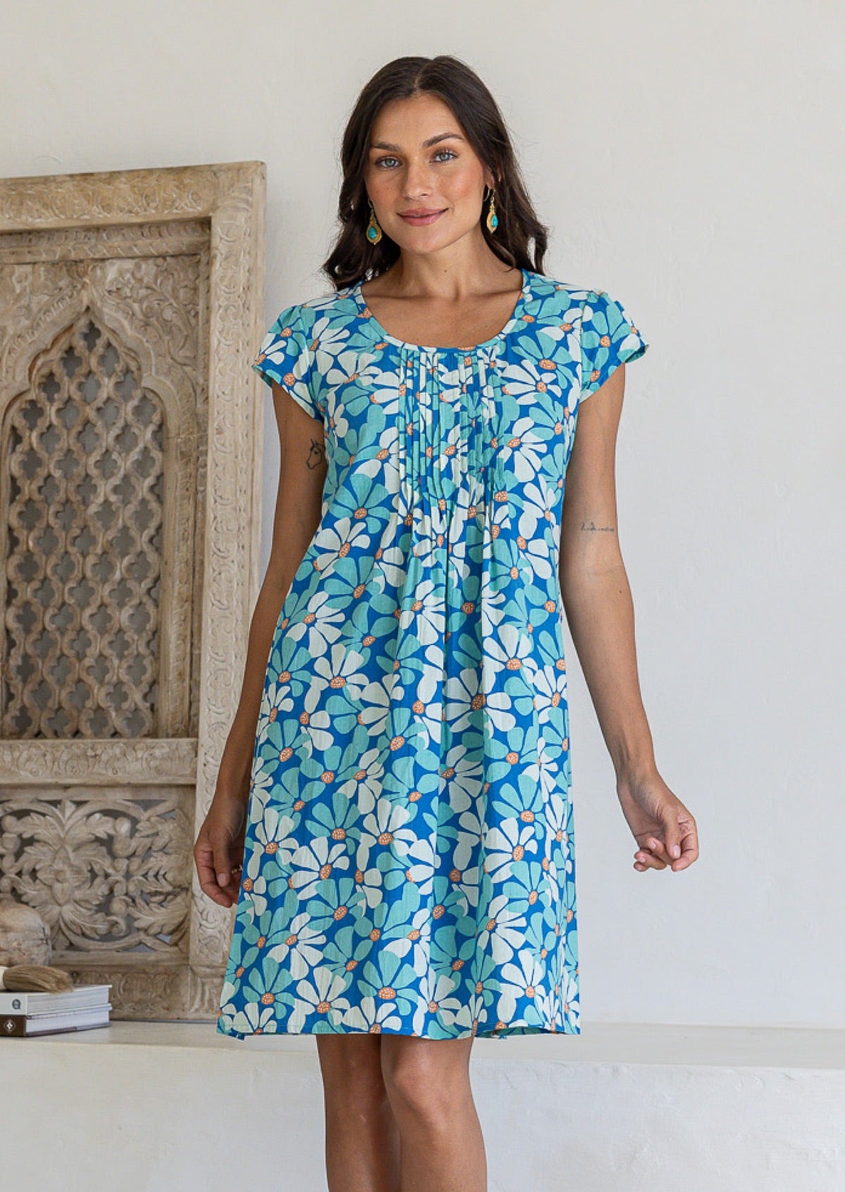 Woman wearing cap sleeved cotton above knee dress in blue and aqua daisy print, with round neckline and pin-tucks across bodice