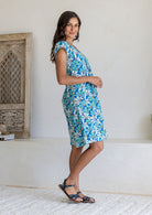 Woman standing side on wearing cap sleeved cotton above knee dress in blue and aqua daisy print, paired with blue sandals
