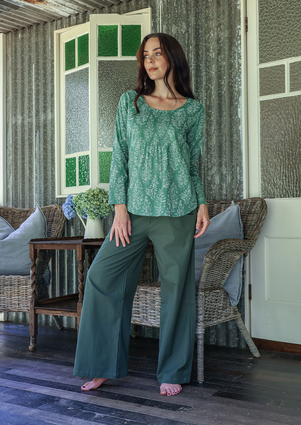 Woman wearing a sage green floral top and pants standing in a room with wicker furniture and a window.