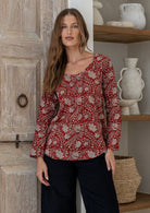 Woman wearing a red 100% cotton floral top standing in a room with wooden door and shelves.