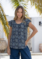 Woman wears navy blue based floral print 100% cotton cap sleeve top with pin tuck detail across the bust and a scoop neckline