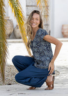 Woman in a blue floral top and navy pants sitting on a wooden floor with palm leaves in the background