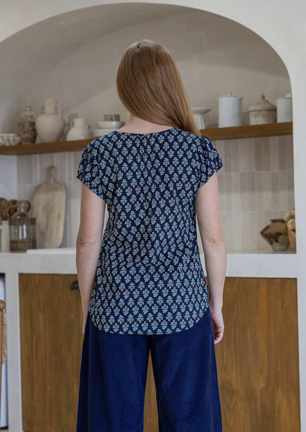 Woman stands backwards wearing  100% cotton top in navy blue based floral print with navy plain pants. 