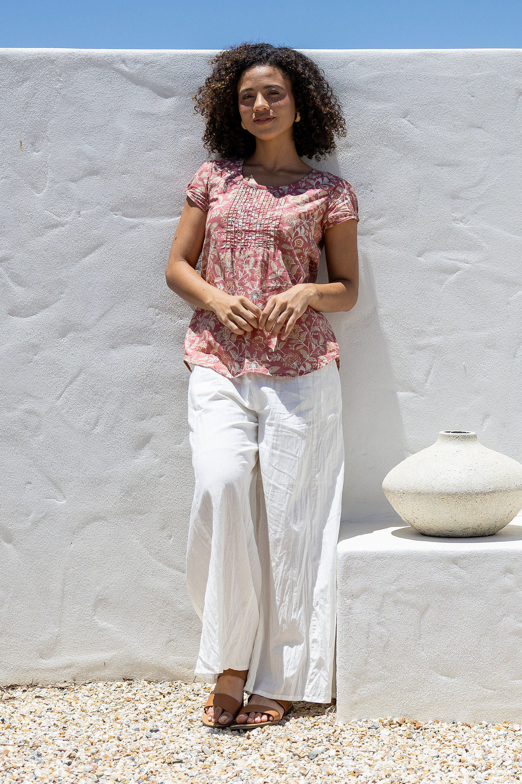 Woman leans on wall wearing 100% cotton top in rose pink floral print with white plain pants. 