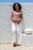 Woman leans on wall wearing 100% cotton top in rose pink floral print with white plain pants. 
