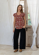 Woman stands wearing  100% cotton top in red based floral print with black plain pants. 
