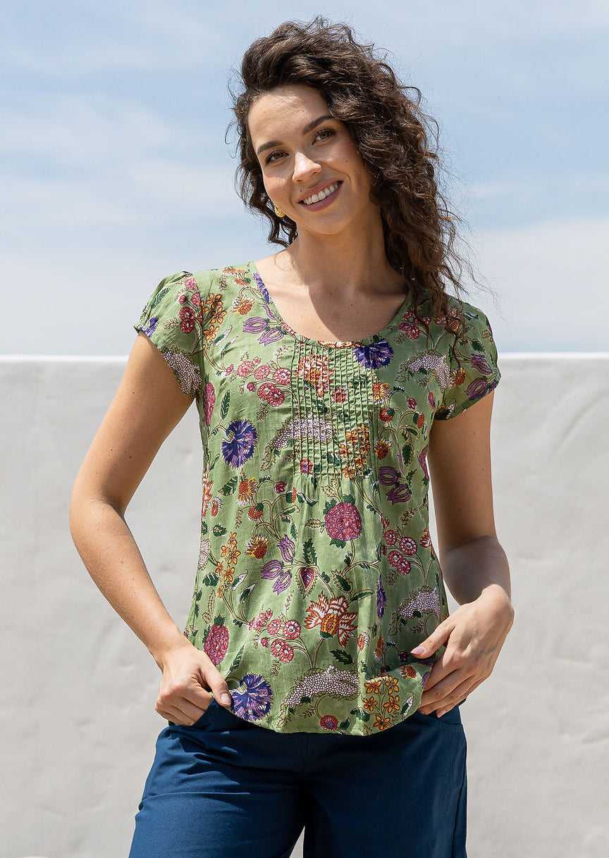 Woman wears moss green base with multi-coloured folk art style florals cotton cap sleeve top with pin tuck detail across the bust and a scoop neckline