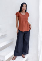 Woman wearing a rust orange cotton top with cap sleeves and pleated detailing, paired with dark cotton pants standing on a white staircase.