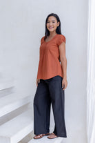 Woman poses in black cotton pants and relaxed fit cotton cap sleeved top in rust orange with pleated detailing across the bodice.