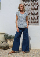 Woman standing with hand in pocket wears 100% cotton short sleeved summer top in white based blue floral print with round neckline and decorative small pleats across the bust.