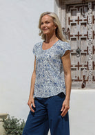 Woman standing near a wooden window wears lightweight cotton top in white based floral print with round neckline, decorative pleats across the bust and cap sleeves.
