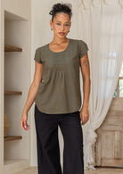 Woman wearing an olive green cotton cap sleeve top and black pants standing in a room with wooden floor and white curtains.